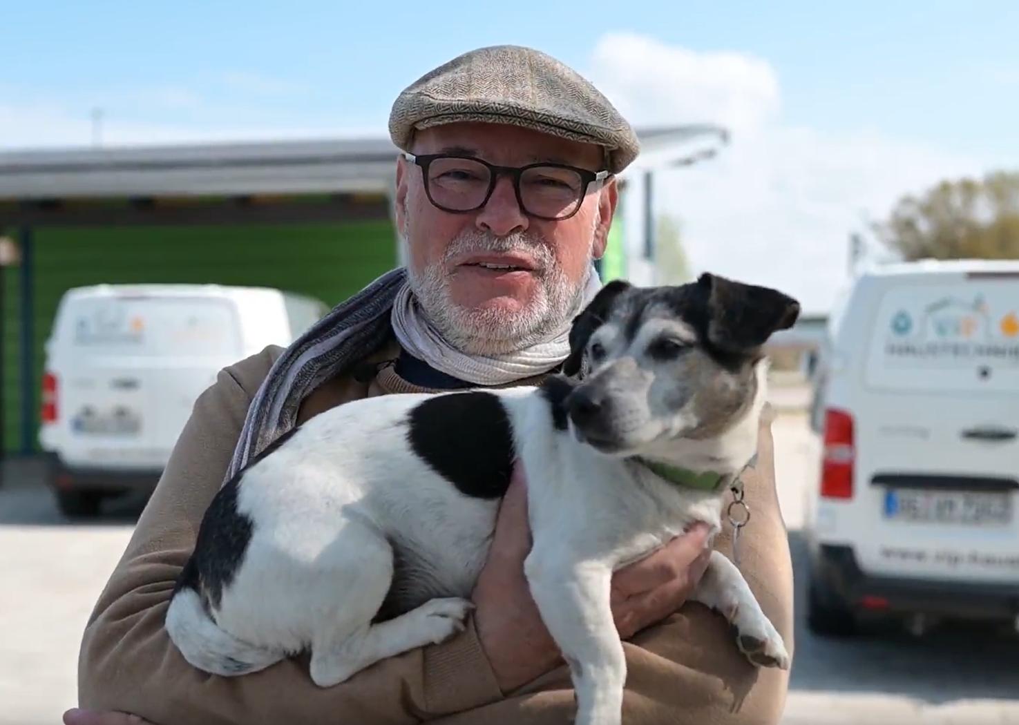 Ein Mann mit Brille und M&uuml;tze h&auml;lt einen kleinen Hund mit schwarz-wei&szlig;em Fell im Arm.